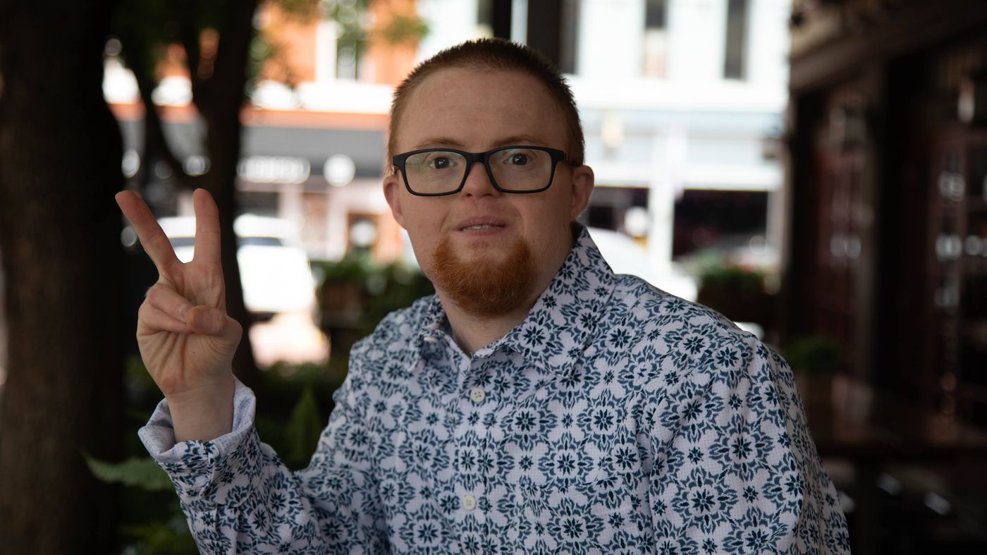 A young man with a red beard and dark-framed glasses, wearing a patterned shirt, smiling at the camera and holding up two fingers in a peace sign.