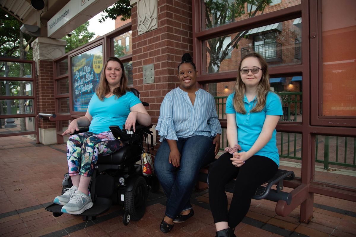 Three young women sitting together at a brick transit station and smiling: one in a power wheelchair, one in a striped blue blouse, and one in a turquoise polo shirt.
