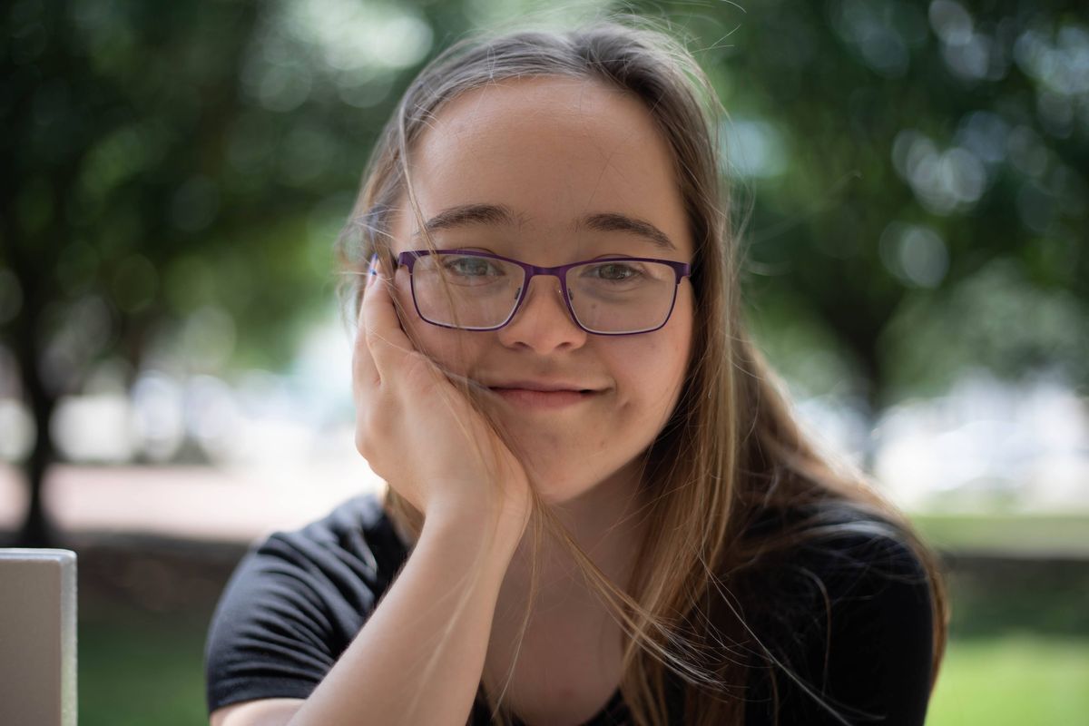 A young woman with long light brown hair and purple glasses, resting her chin on her hand and smiling at the camera, with green trees behind her.